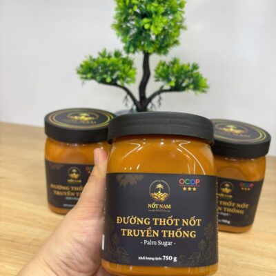 Tay cầm hũ đường thốt nốt sệt Nốt Nam 750g, phía sau là các hũ cùng loại và chậu cây xanh